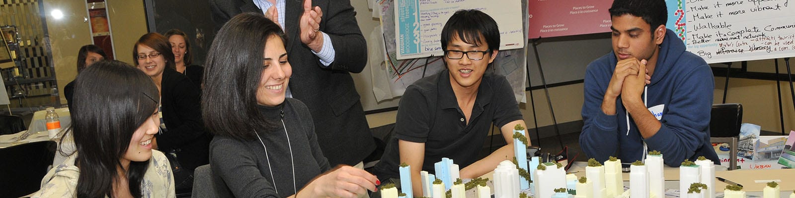 Photo of people sitting around a table with a model at the centre and pointing to a building.