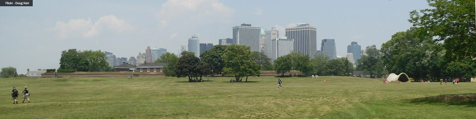 Photo of people walking through a large green space with tall buildings in the background.